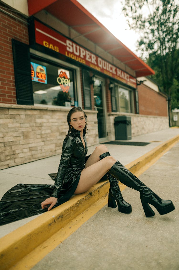 Young Brunette Woman In Black PVC Raincoat And High-Heeled Platform Boots Sitting On A Curb