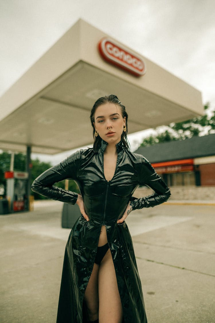 Young Brunette Woman In Black PVC Matrix Style Trench Coat Posing At A Gas Station 