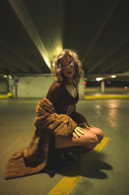 Fashionable woman in a coat poses in a dimly-lit urban parking garage, exuding confidence and style.