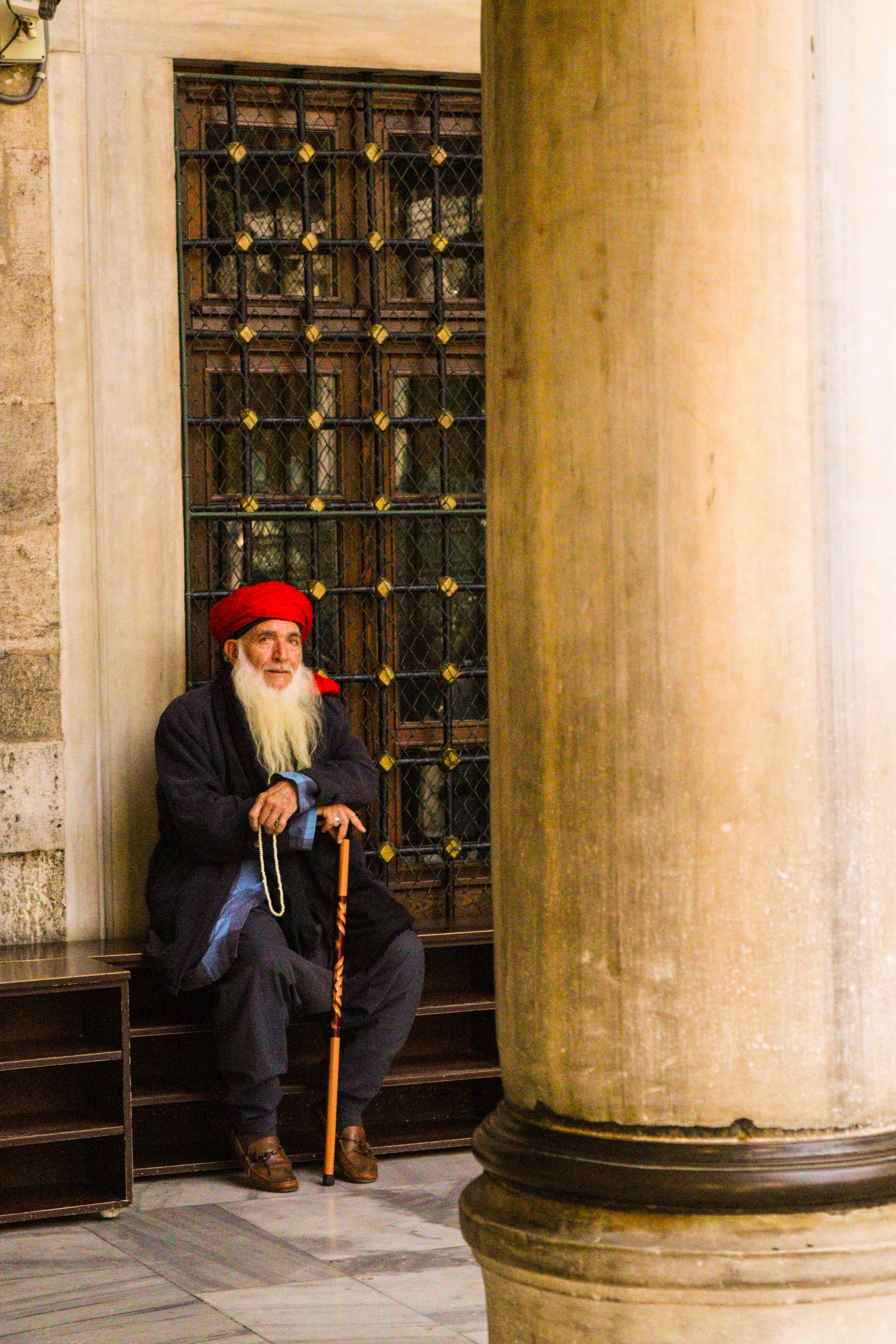 Elderly man with a long beard and red hat sitting by a mosque gate, holding a cane.