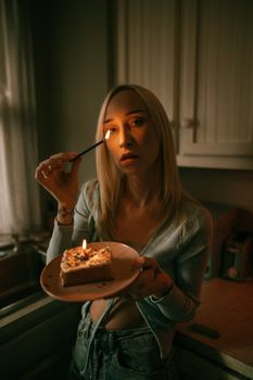 A woman holding a cake with a lit candle in a dimly lit kitchen, creating a moody ambiance.
