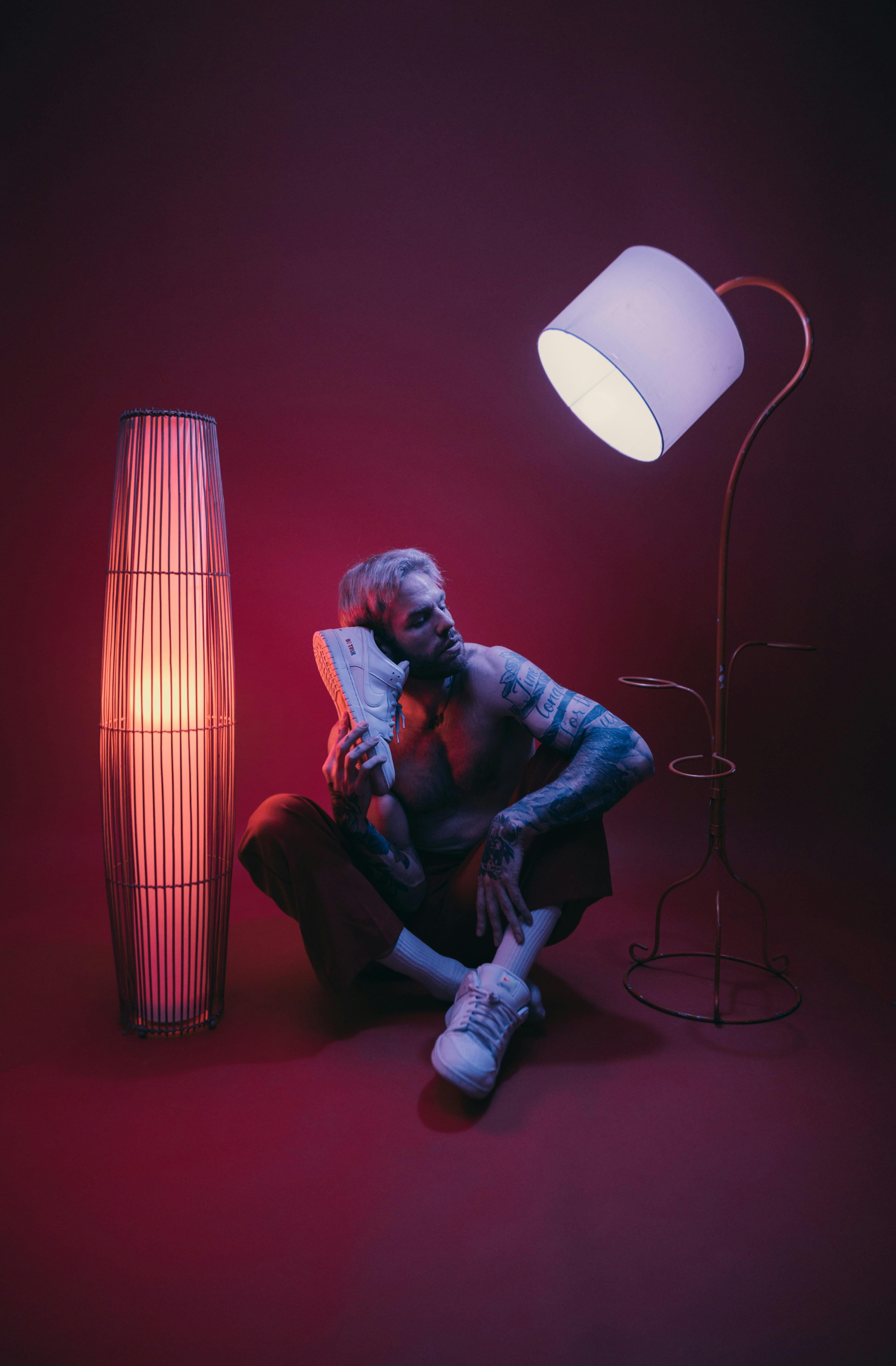 Tattooed man posing with sneakers under artistic lighting in modern studio.