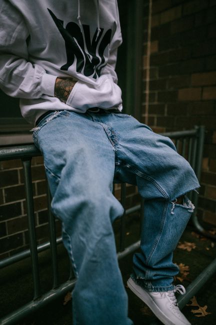 Close-up of a person in ripped jeans and hoodie sitting on an urban railing.
