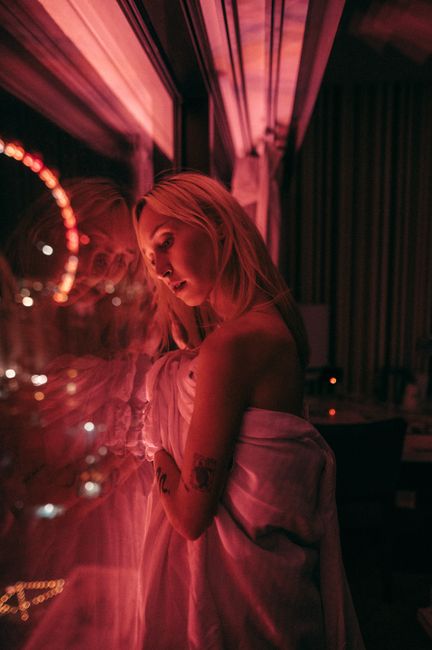 A woman wrapped in a sheet gazes thoughtfully at a window reflecting pink neon light.