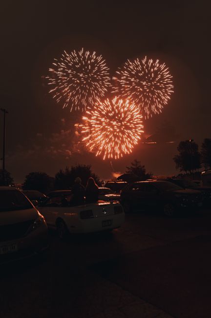 A couple watches fireworks from a convertible in a parking lot, capturing a festive night scene with urban vibes.