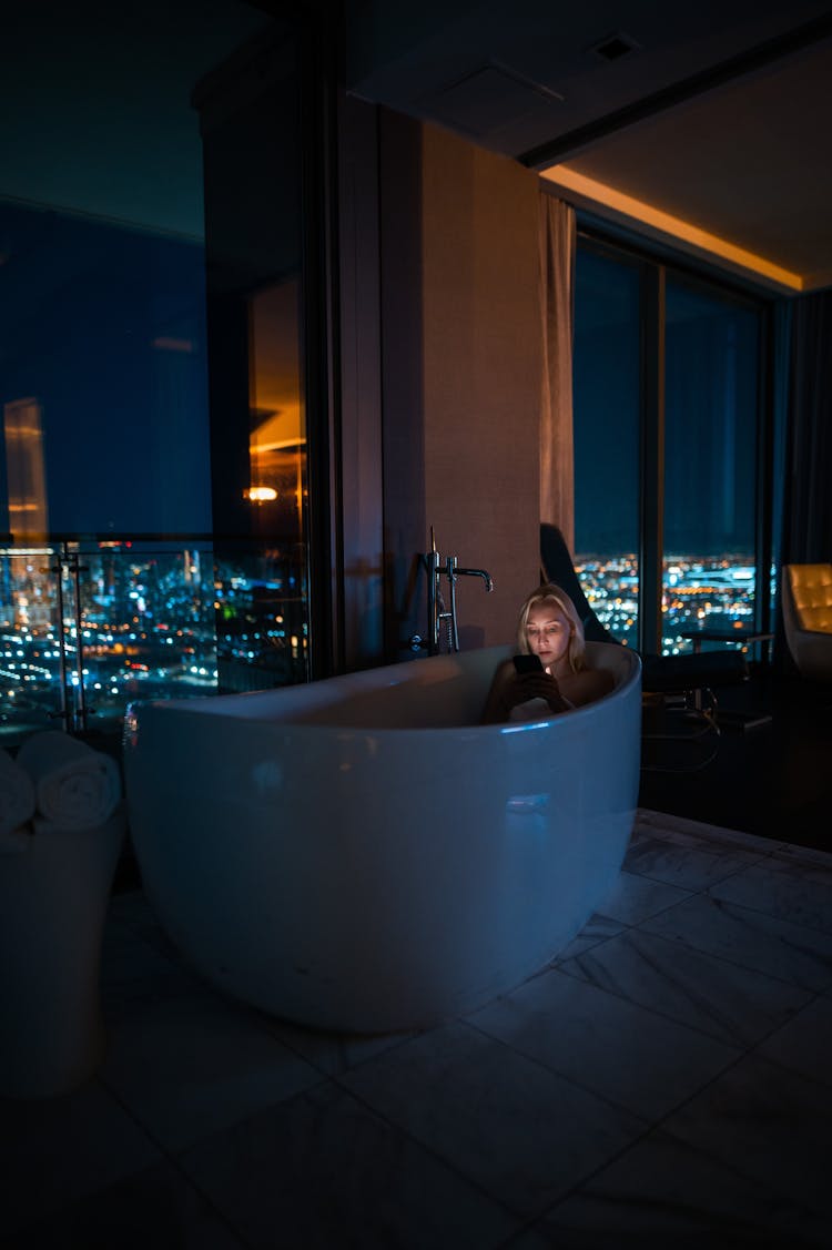 Woman In Bathtub Near Windows With City Lights Behind