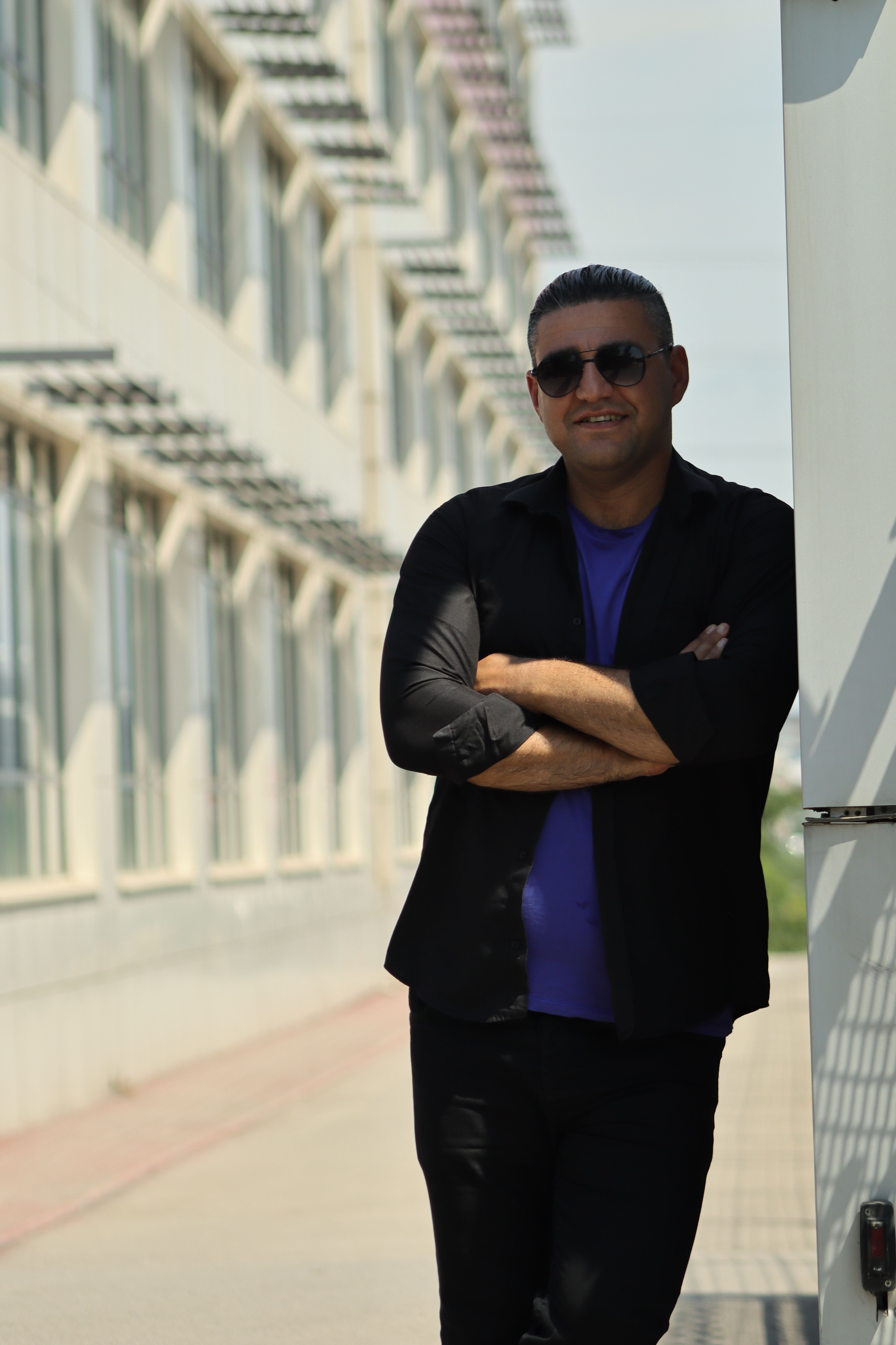 Fashionable man in sunglasses wearing black clothes, leaning against a wall in an urban setting.