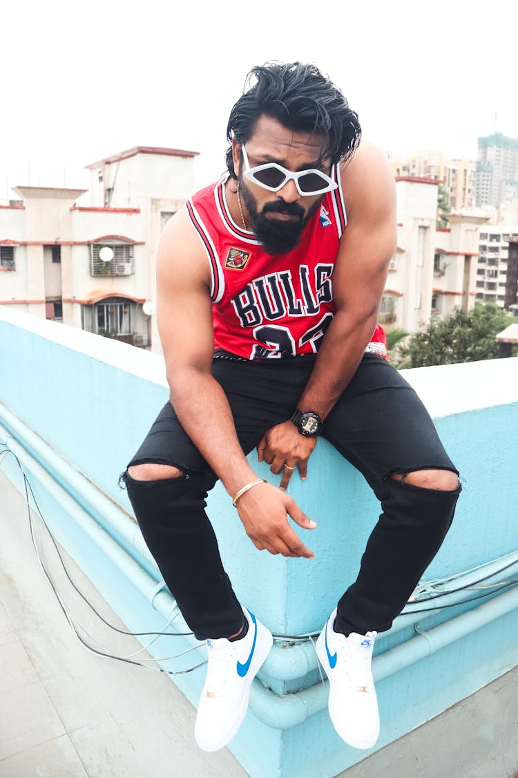 Brunette Man In Ripped Skinny Jeans And Red Basketball Jersey Sitting On A Parapet