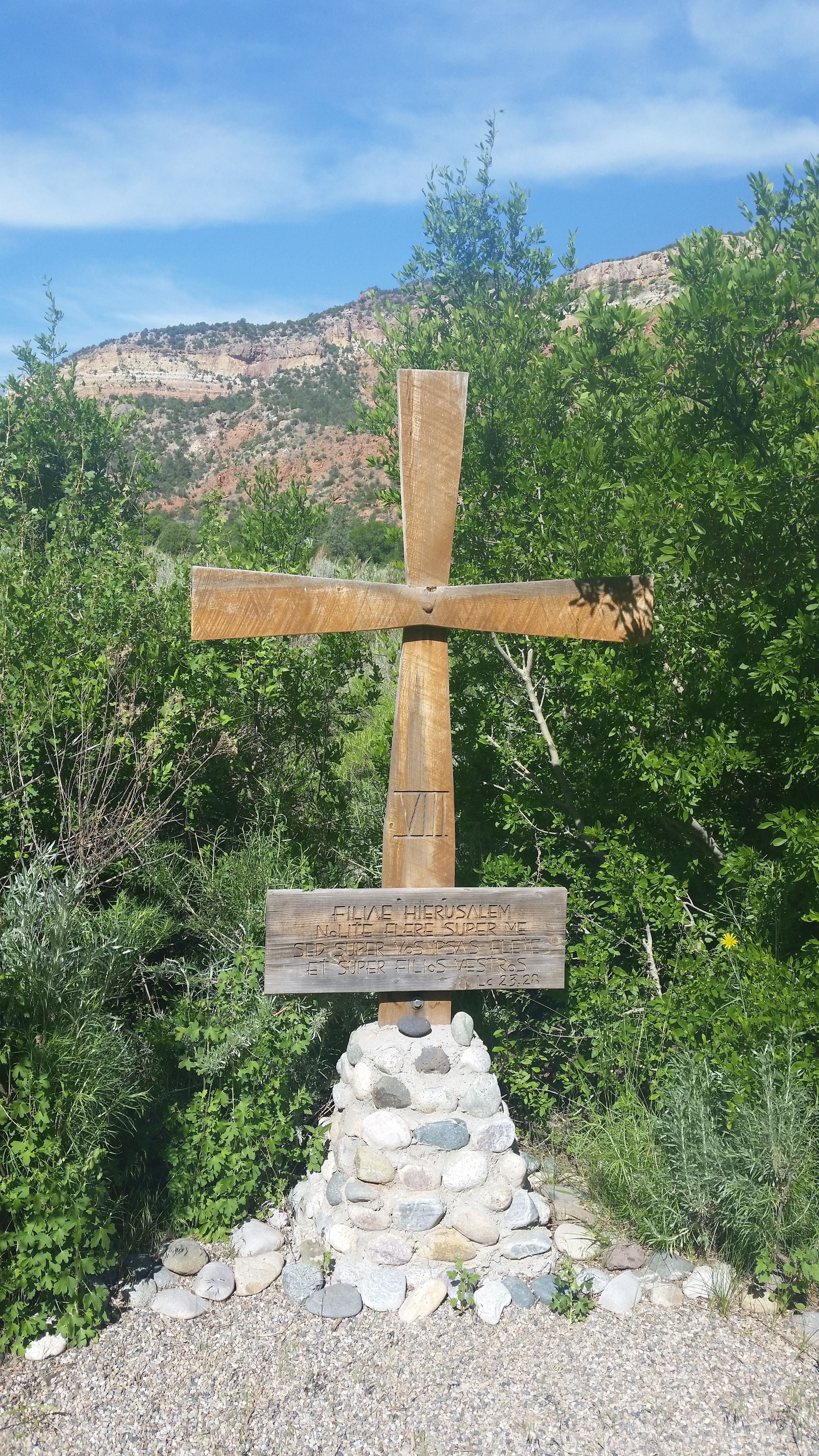 A wooden cross stands amidst lush greenery with mountains in the background.