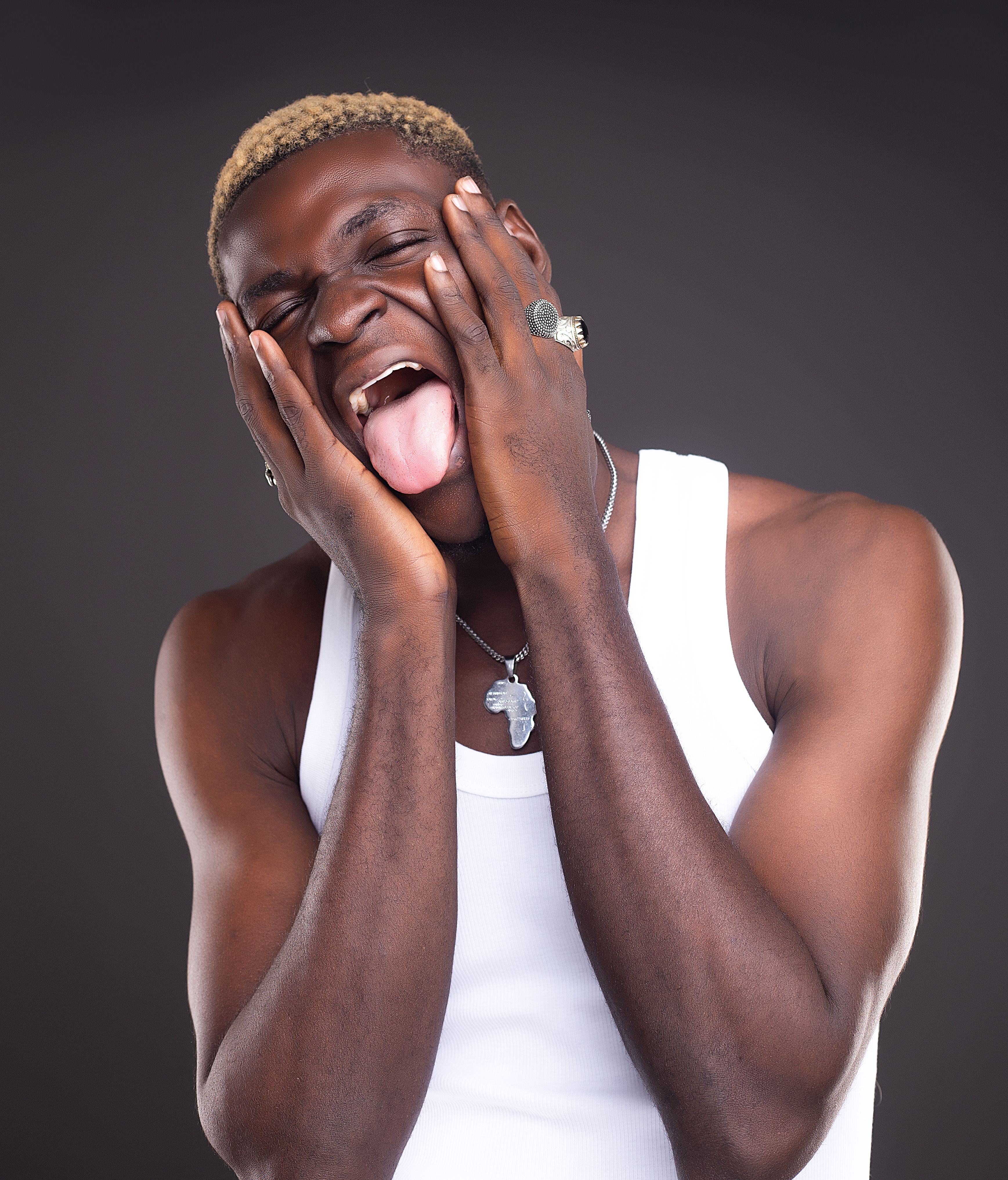 A man in a white tank top showing expression with his tongue out, hands on face, in a studio setting.