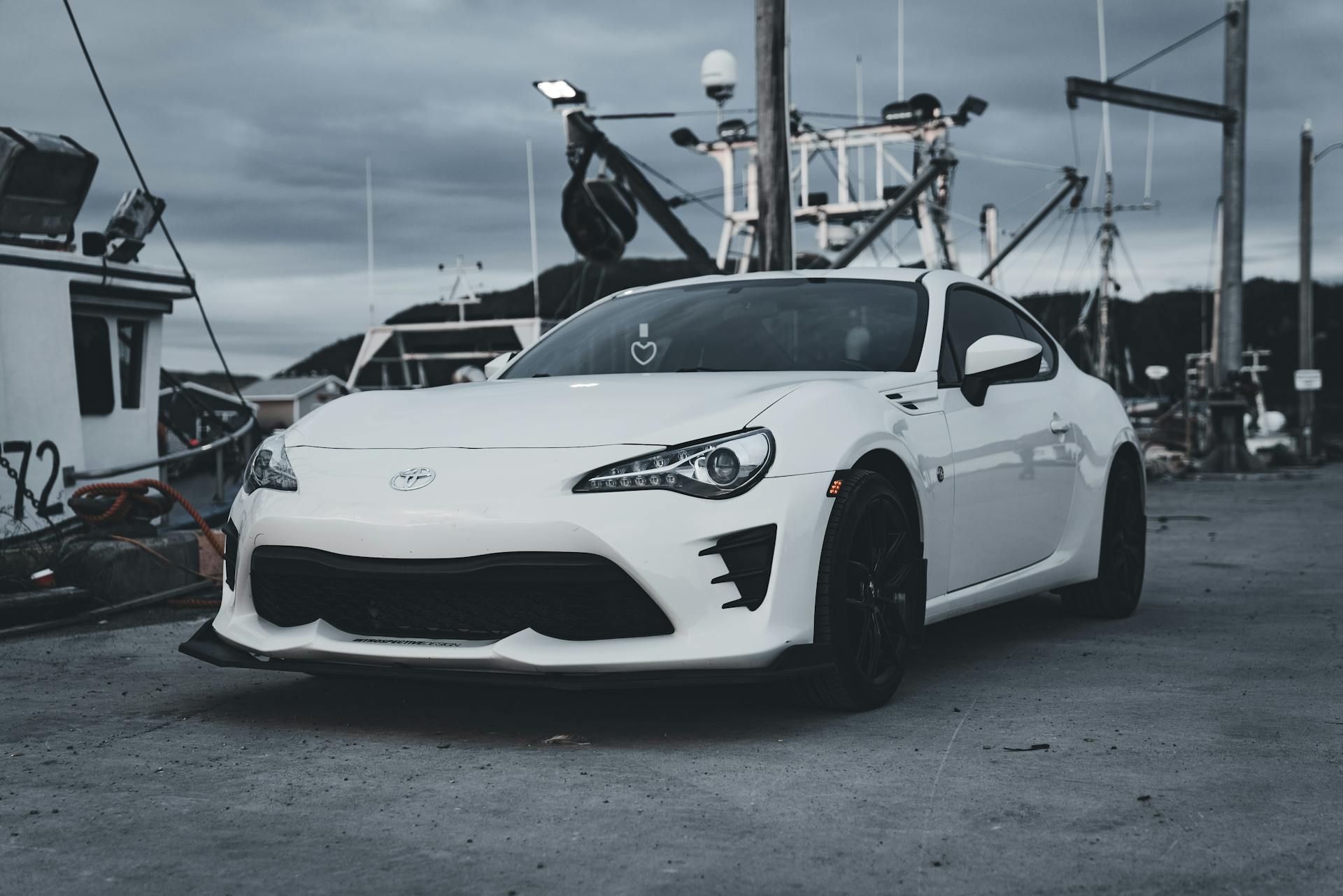 A sleek white Toyota GT86 parked in a scenic harbor with boats in the background.