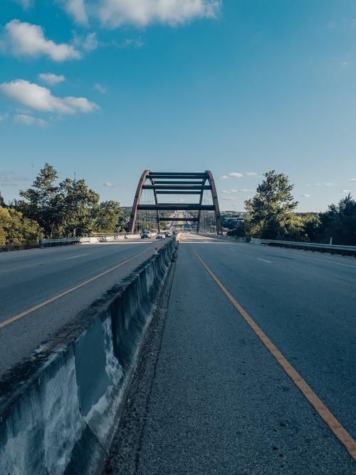 Free Peaceful highway road leading to an arch bridge surrounded by greenery under a clear blue sky. Stock Photo