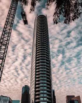 低角度拍摄的一座现代摩天大楼，顶部装有起重机，映衬着多云的天空，展现了城市建筑的景象