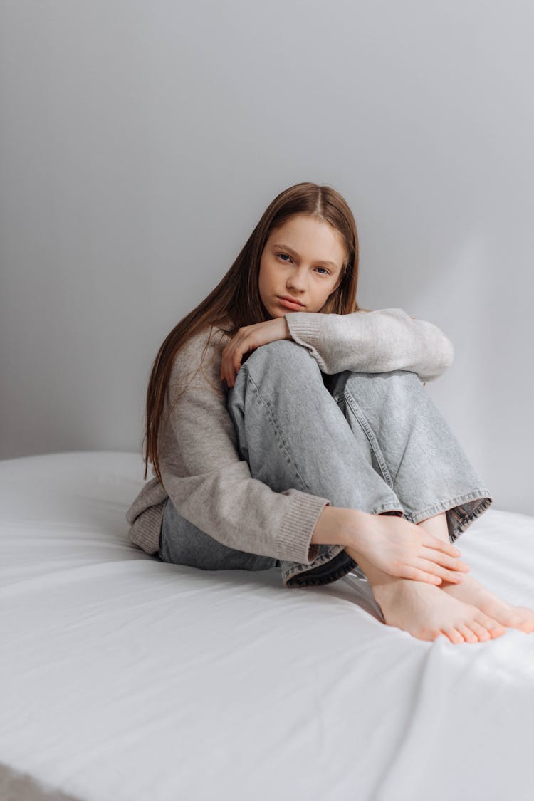 Teenager Girl In A Grey Jumper And Jeans Sitting On A Bed