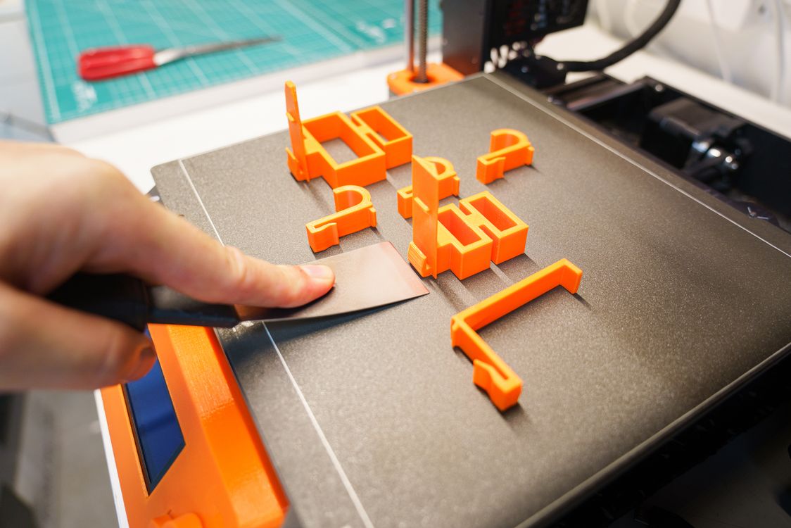 Hand carefully removing a freshly 3D printed orange object from the printer bed using a spatula