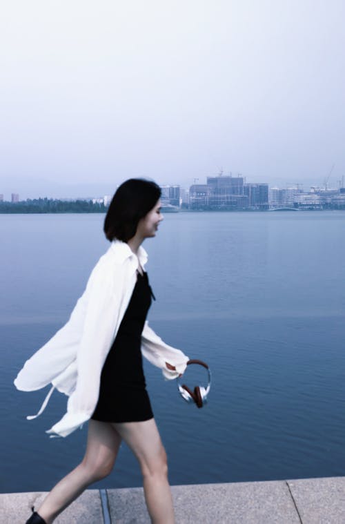 Female Model Wearing a White Shirt and a Black Dress Walking along the Coast with Headphones in Hand