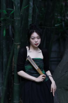 Asian woman in green dress holding a fan, standing in a bamboo forest, serene and elegant.