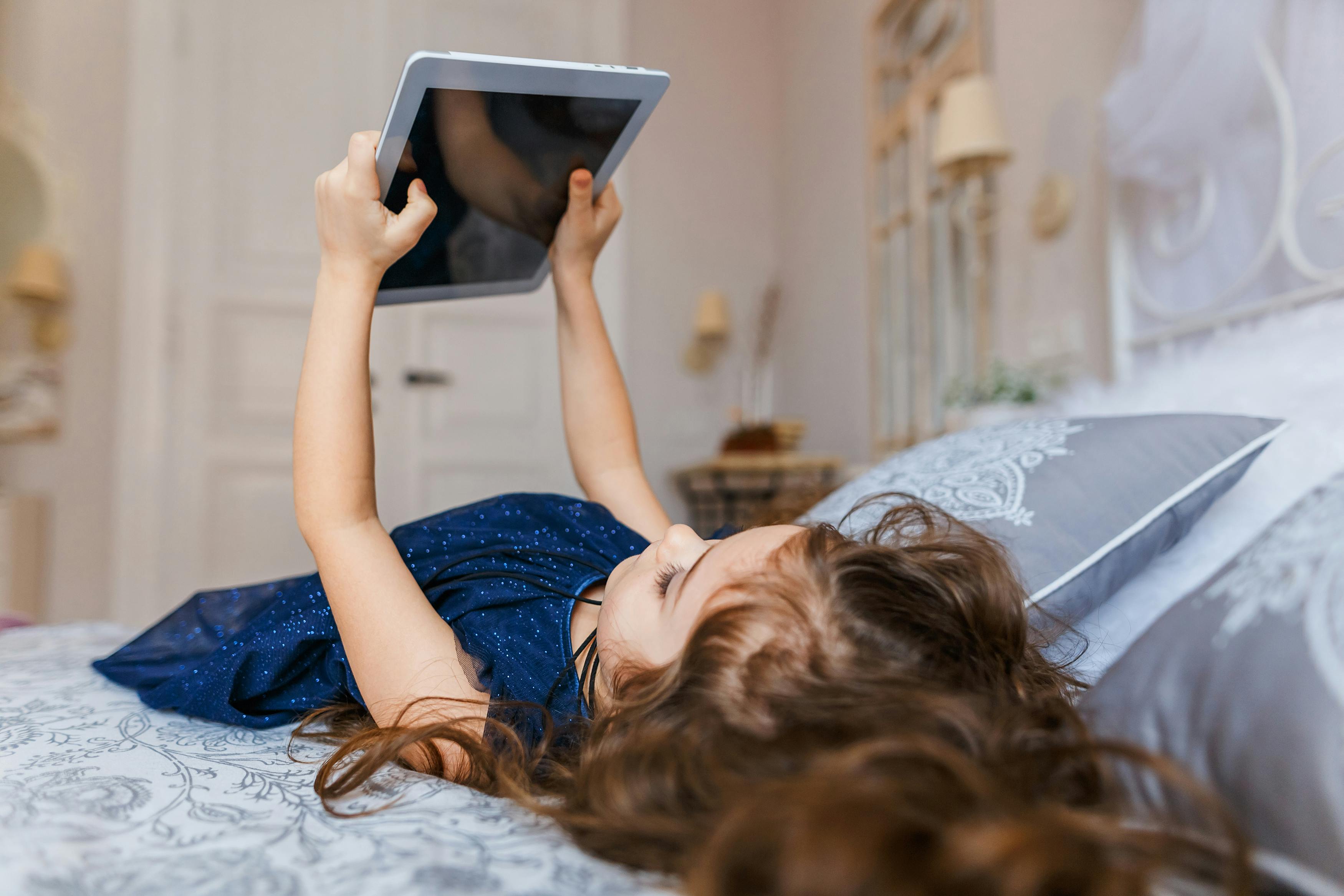 cute little girl with curly hair using digital tablet while lying in bed