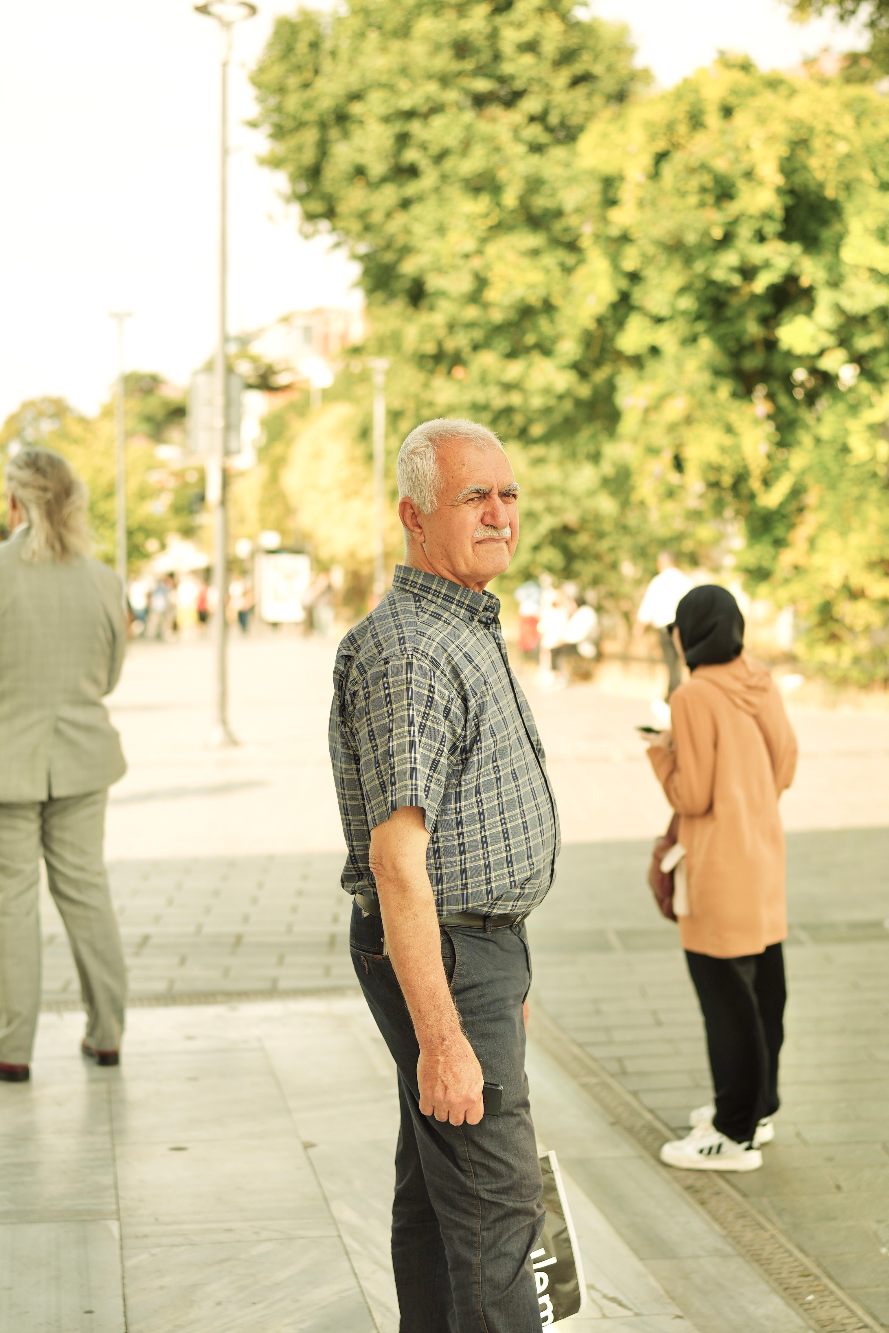 Senior man in casual attire stands on a busy city sidewalk with trees in the background.