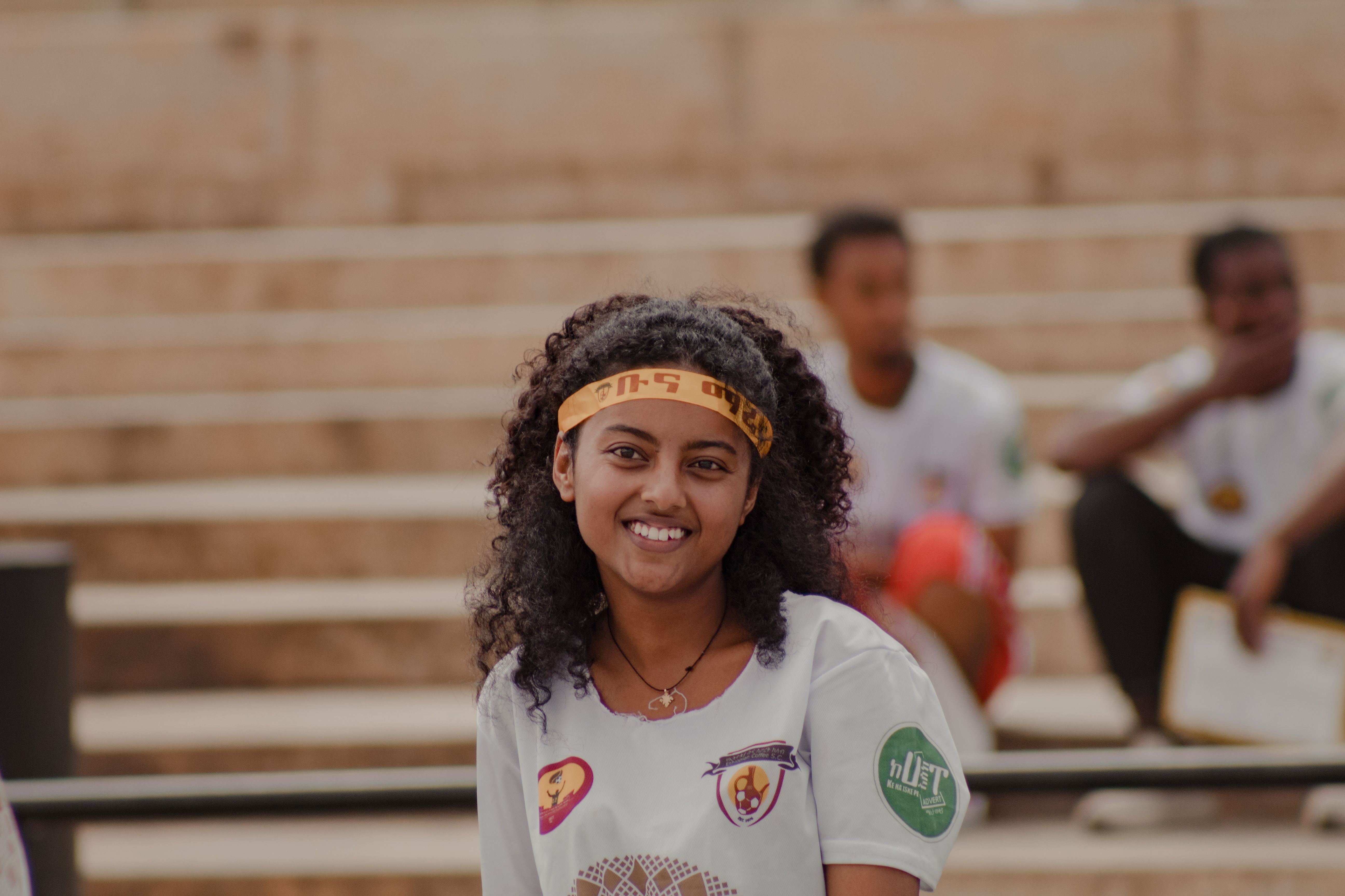 Portrait of a smiling woman with curly hair and a headband, sitting outdoors with a blurred background of people.