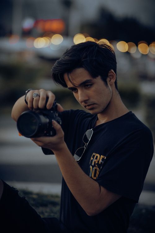 Free Young male photographer focusing his camera outdoors during twilight in an urban setting. Stock Photo