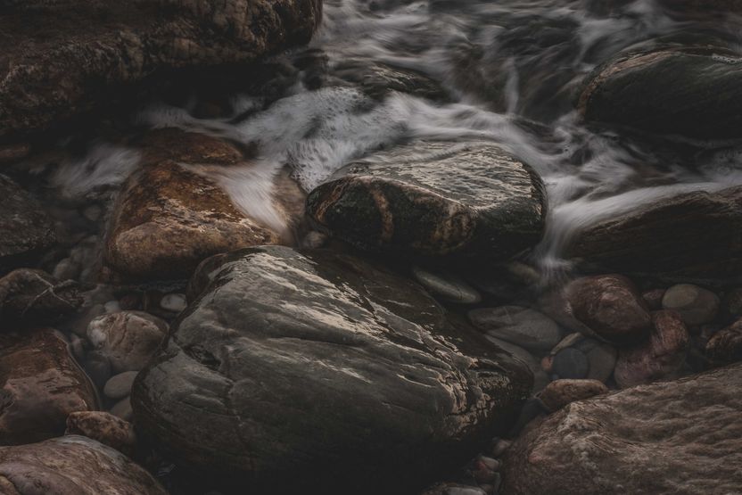 A long-exposure close view of a shallow stream running over large wet stones, with one dark boulder sharp at the centre and the water blurred smooth around it.