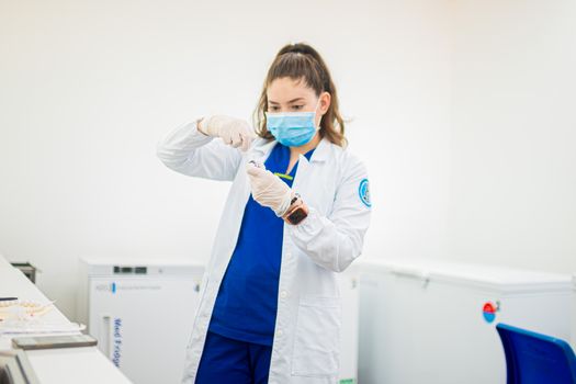 A female healthcare worker in uniform and mask prepares a syringe in a medical facility.
