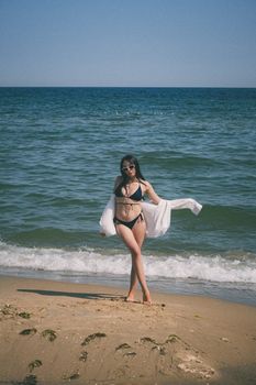 A fashionable woman in a black bikini enjoys a sunny day at the beach, posing by the ocean.