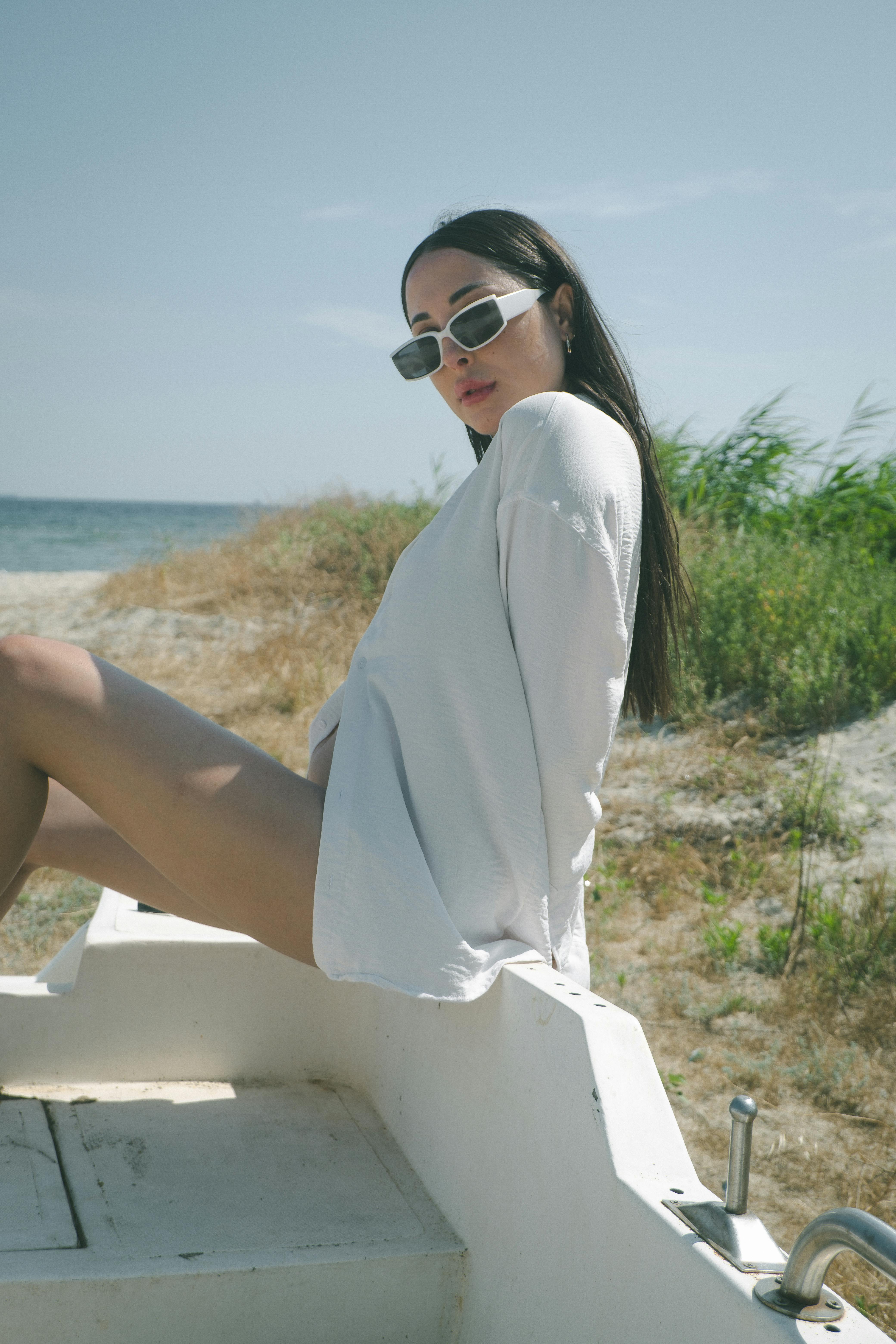 Free Woman in sunglasses and white top posing on a beach chair by the shore, exuding relaxed elegance. Stock Photo