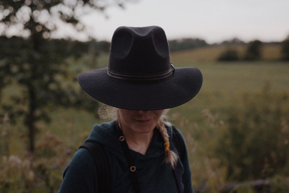 A woman wearing a cowboy hat walks in a serene countryside setting under natural daylight.