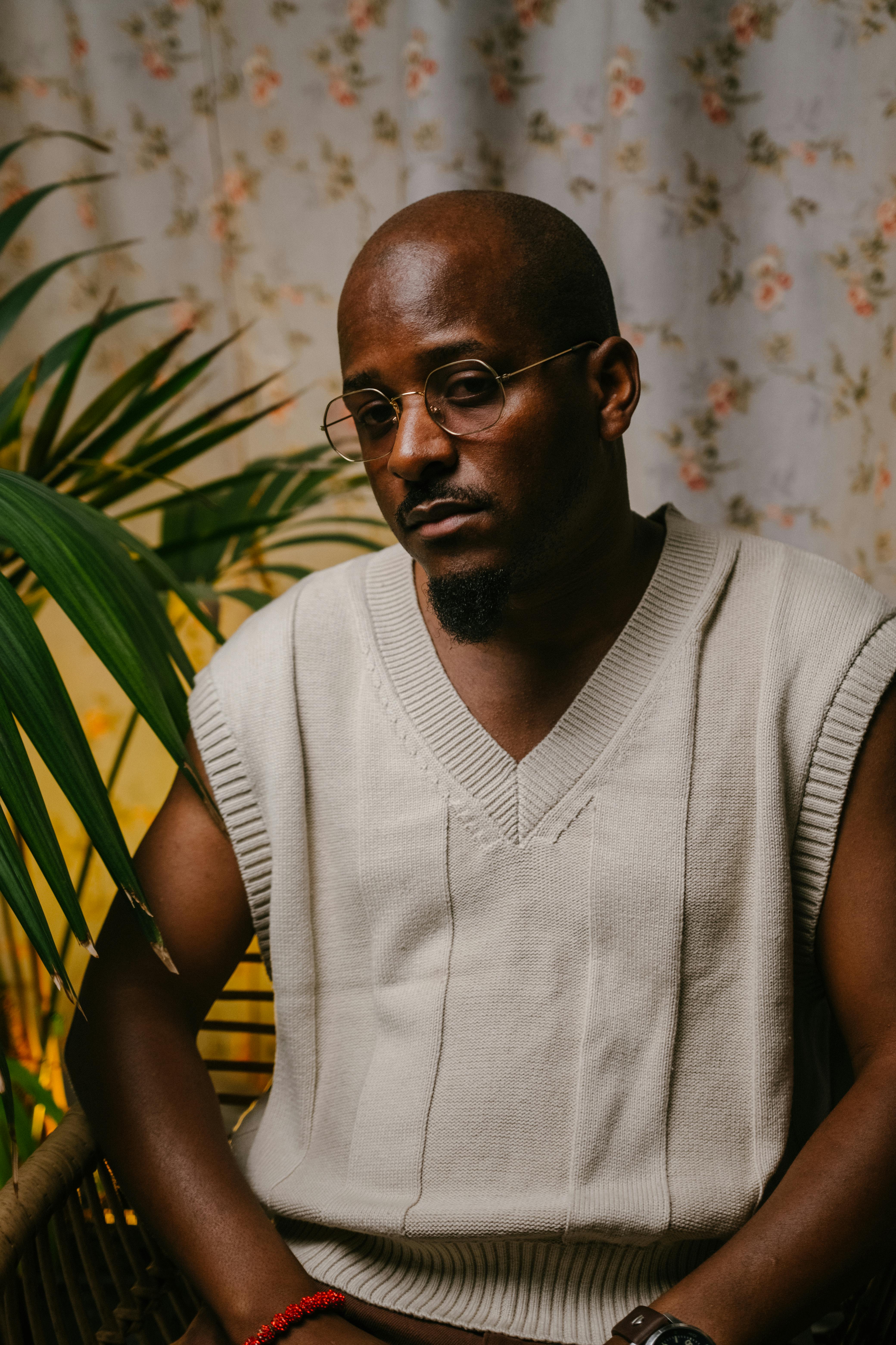 Portrait of a fashionable man wearing a sleeveless sweater, sitting indoors with a potted plant backdrop.