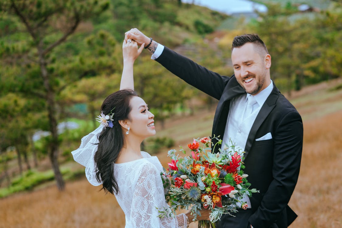 A joyous newlywed couple dancing outdoors, with the bride twirling under the groom's arm. The bride, in a lace wedding dress and a floral hairpiece, beams with happiness as she holds a vibrant bouquet of red and orange flowers. The groom, in a classic black suit and tie, looks at her affectionately. They are surrounded by a natural landscape of green hills and trees, under a soft, overcast sky.