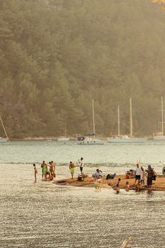 People enjoy a sunny day by the beach with sailboats and forest in the background.