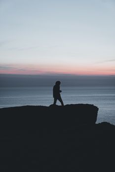 A solitary figure walks along a cliff at sunset, silhouetted against the colorful horizon.