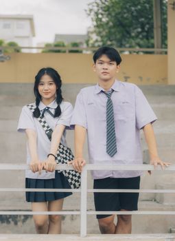 Two young Asian people smiling, wearing school uniforms, leaning on a railing outdoors.
