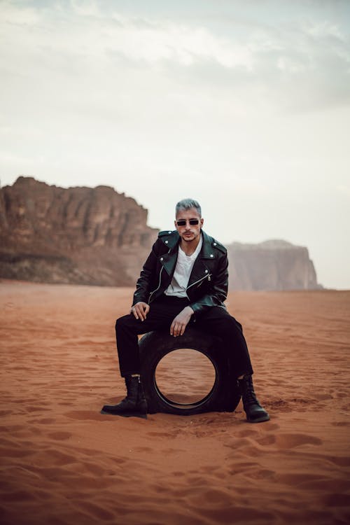 Man in Black Jacket Sitting on Tire on Desert