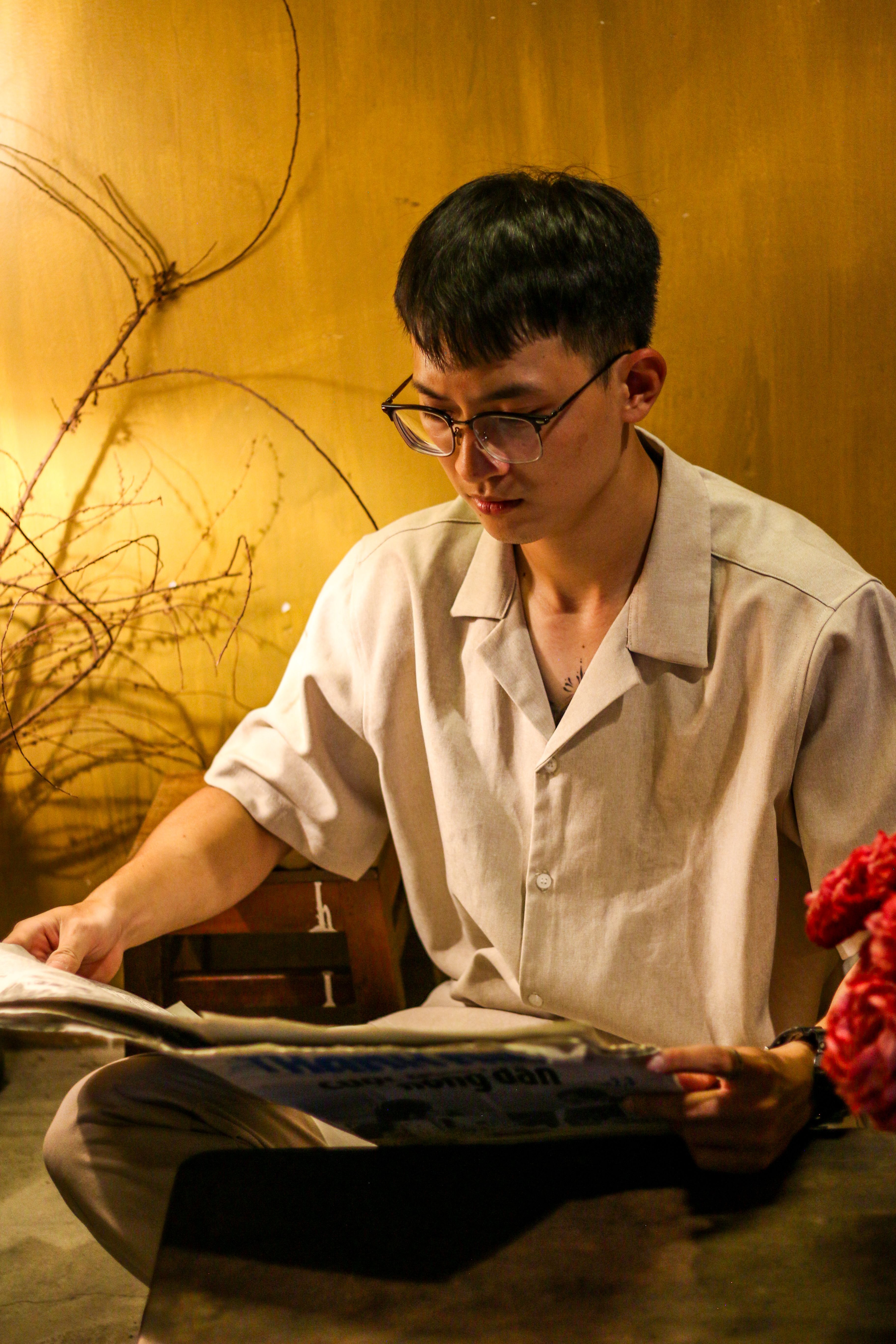 A young man wearing glasses reads a newspaper indoors under warm lighting.