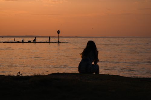 Free A woman sitting by the sea, silhouetted against a vibrant sunset sky, reflecting tranquility. Stock Photo