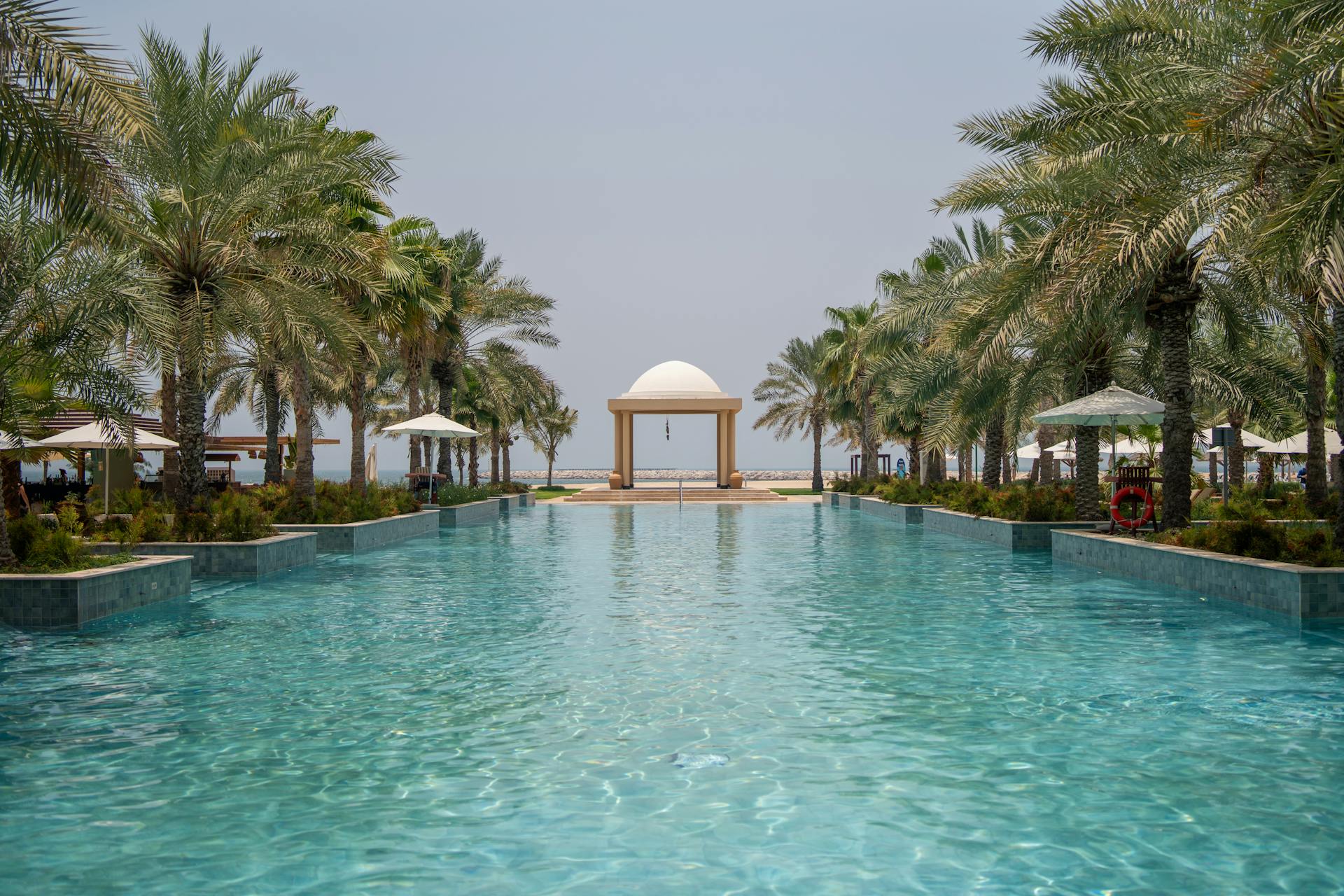 A serene luxury resort pool surrounded by palm trees and a central gazebo under a clear sky.