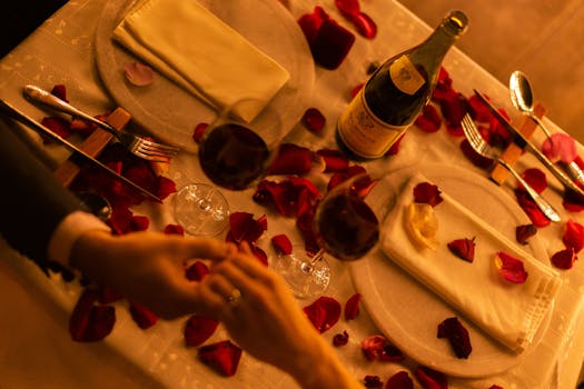 Intimate dinner setting with red wine, rose petals, and a couple holding hands.