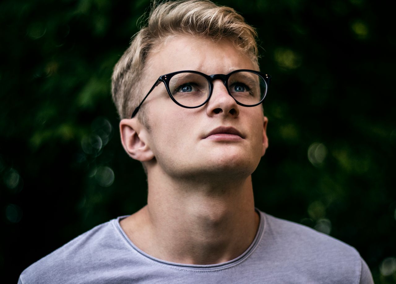 a thoughtful young man wearing eyeglasses looks upwards in an outdoor
