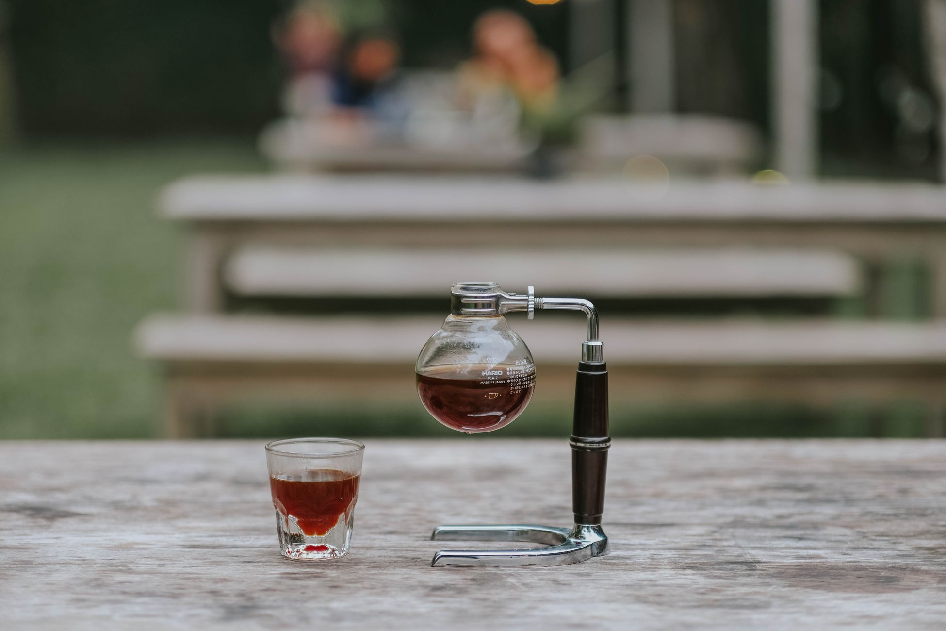 Close-up of a siphon coffee maker and coffee cup on a wooden table outdoors.