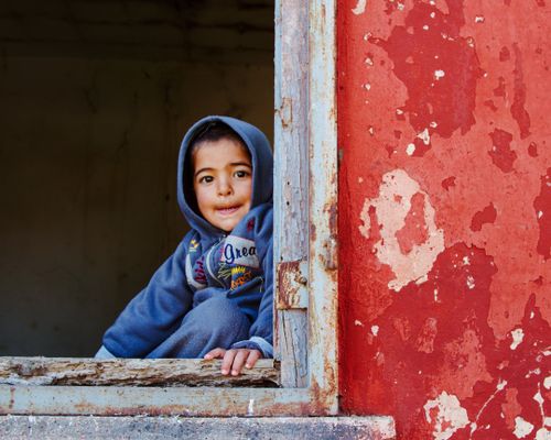 Free Smiling child in a blue hoodie sitting in a rustic window frame of an abandoned building. Stock Photo