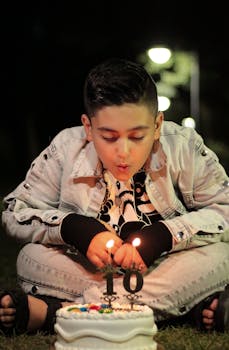 A boy sits on grass, blowing candles on a birthday cake, capturing a joyful moment at night.