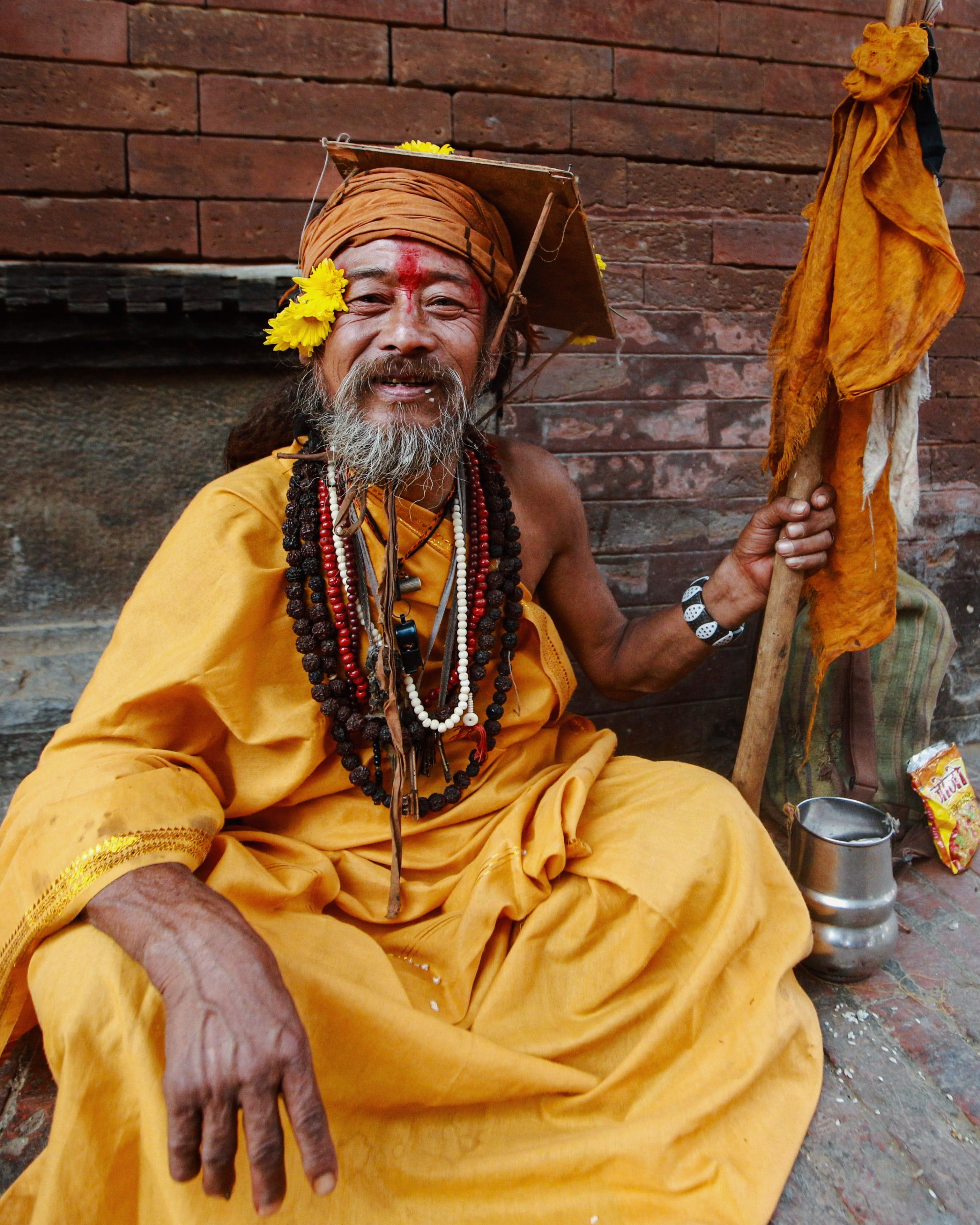 A cheerful sadhu dressed in orange with flowers, sitting against a wall, showcasing spiritual serenity.