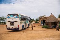 A white bus stops at a rural village with a tribal hut and local restaurant.