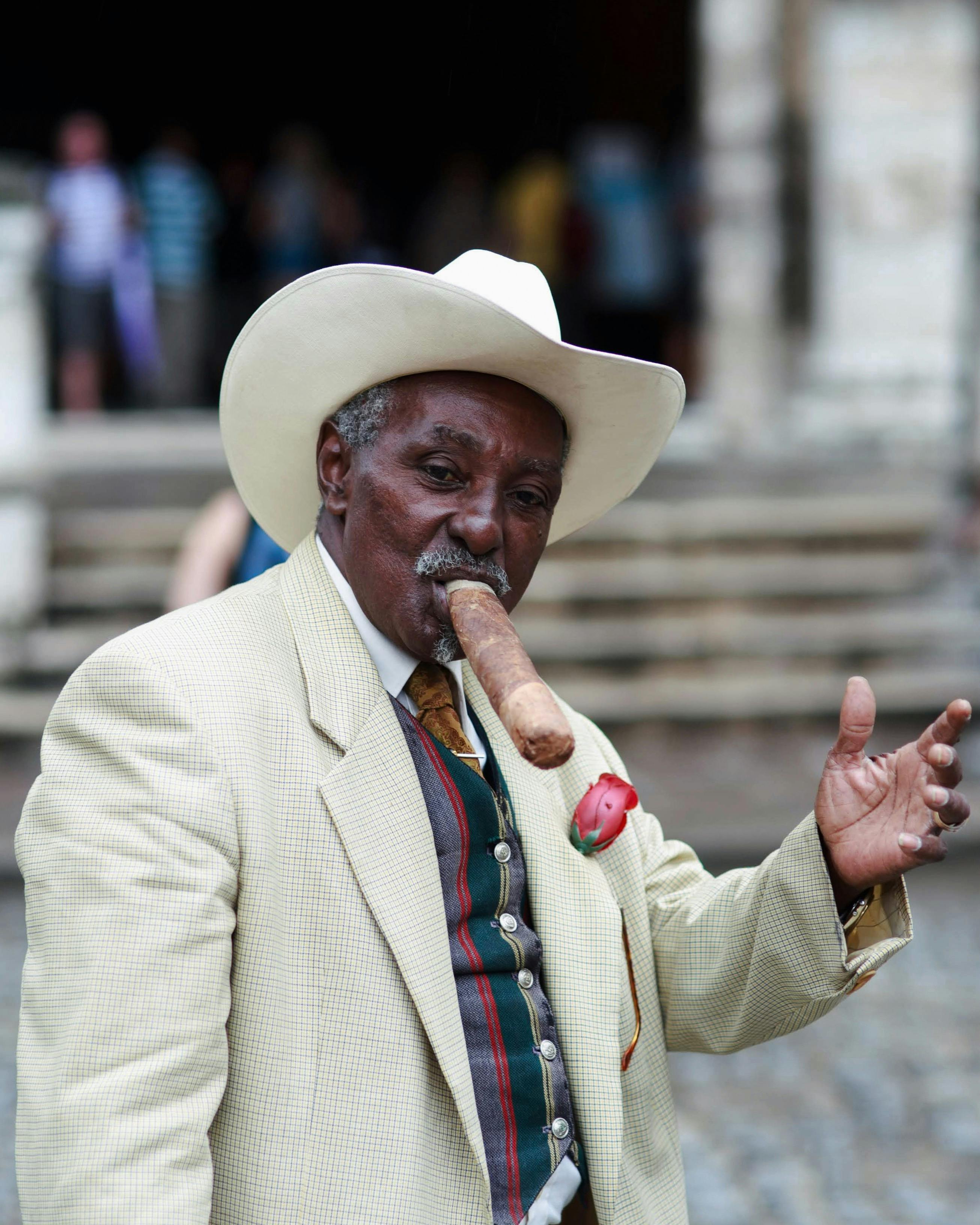 Elderly man in suit and hat enjoying a cigar with a confident pose outdoors.