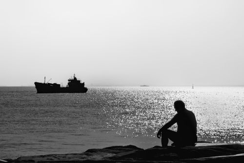 Free Black and white silhouette of a man sitting by the sea with a ship in the distance. Stock Photo