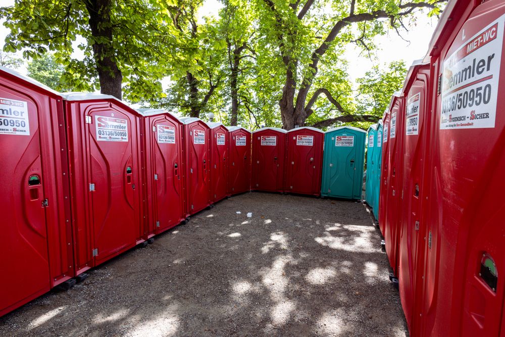 Row of red standard portable toilets set up outdoors under trees