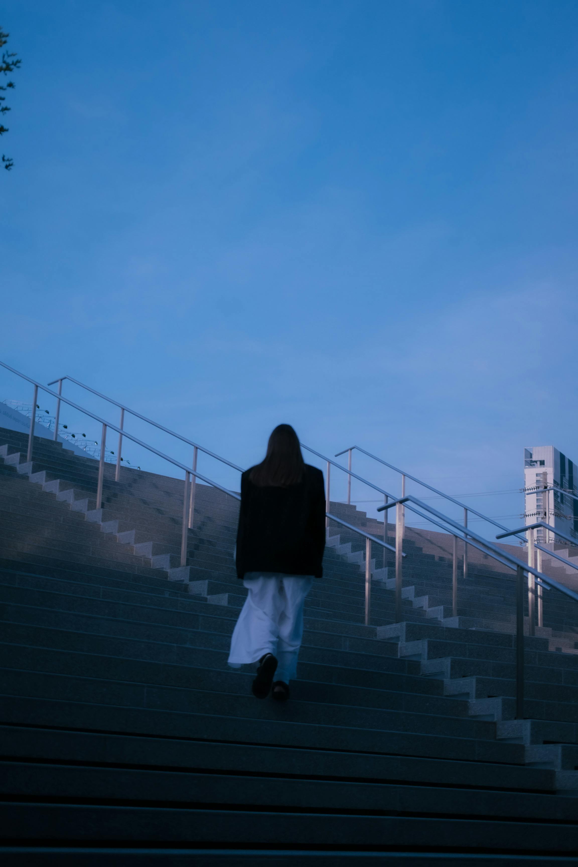 A woman in black and white attire ascends stairs at twilight in Bangkok. Modern architecture visible.